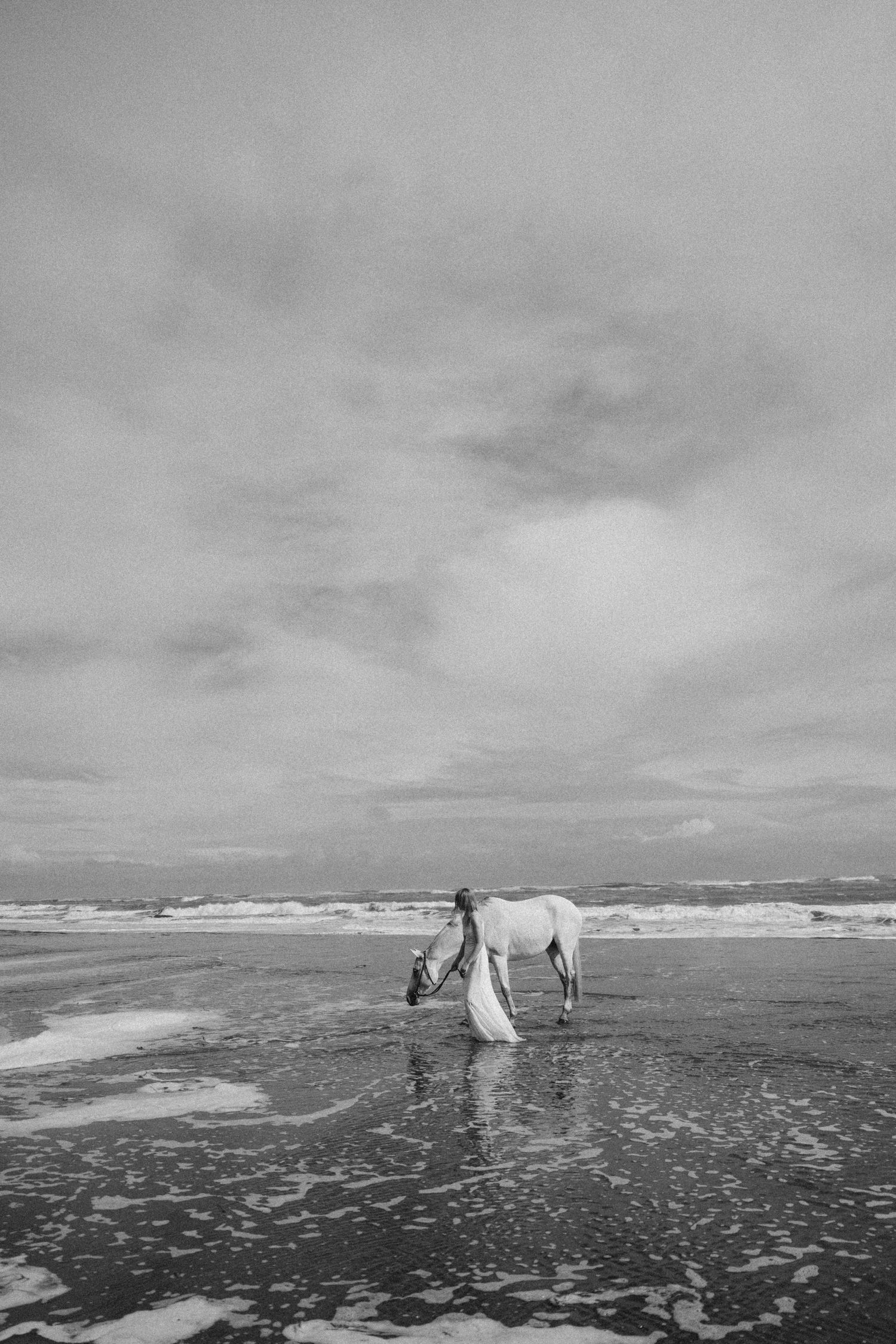 Black and white photo of a person in a white dress walking a white horse on a misty beach with dramatic cloudy skies.