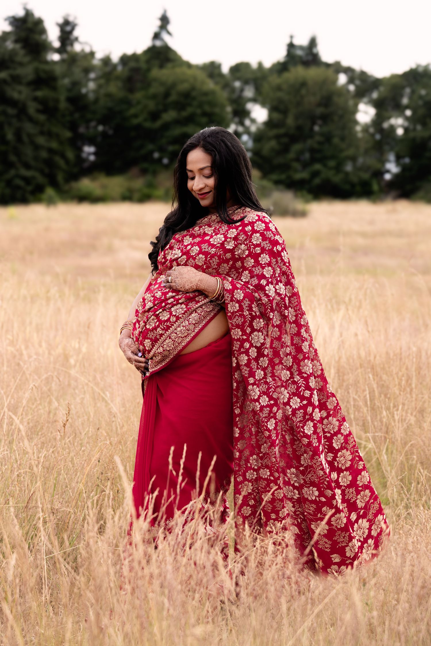 Pregnant woman in red floral saree standing in golden field, cradling baby bump, trees in background.