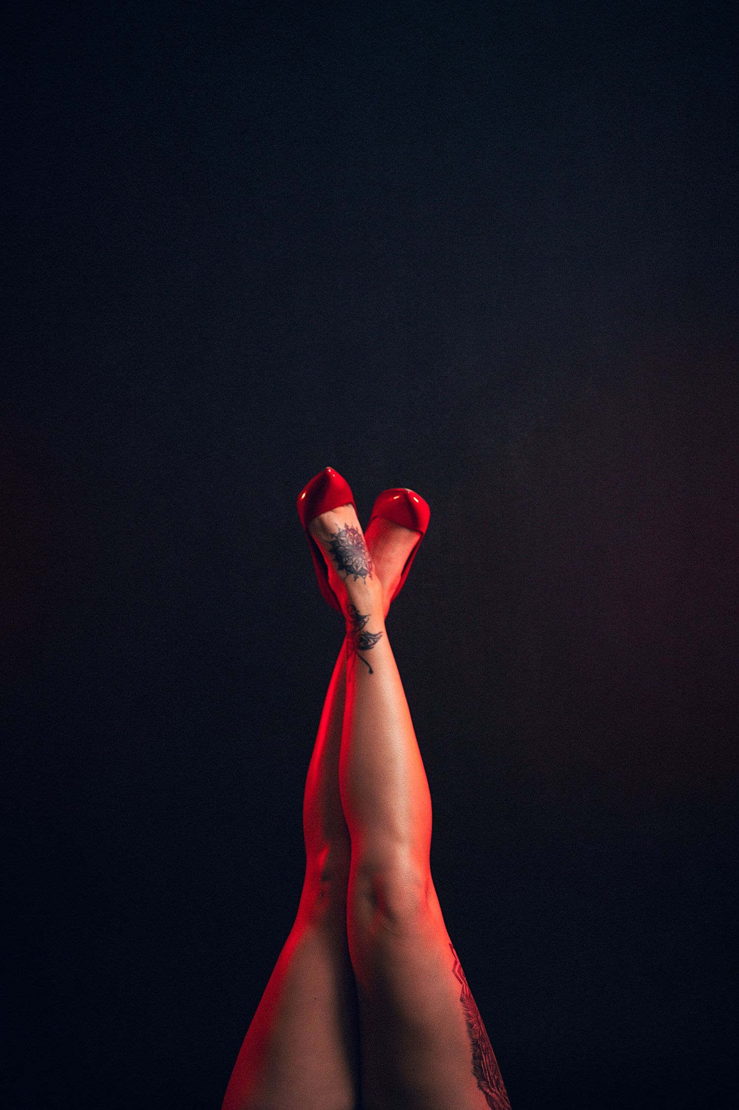 Woman's legs raised up wearing red high heels and fishnet stockings with a tattoo on ankle against dark background.