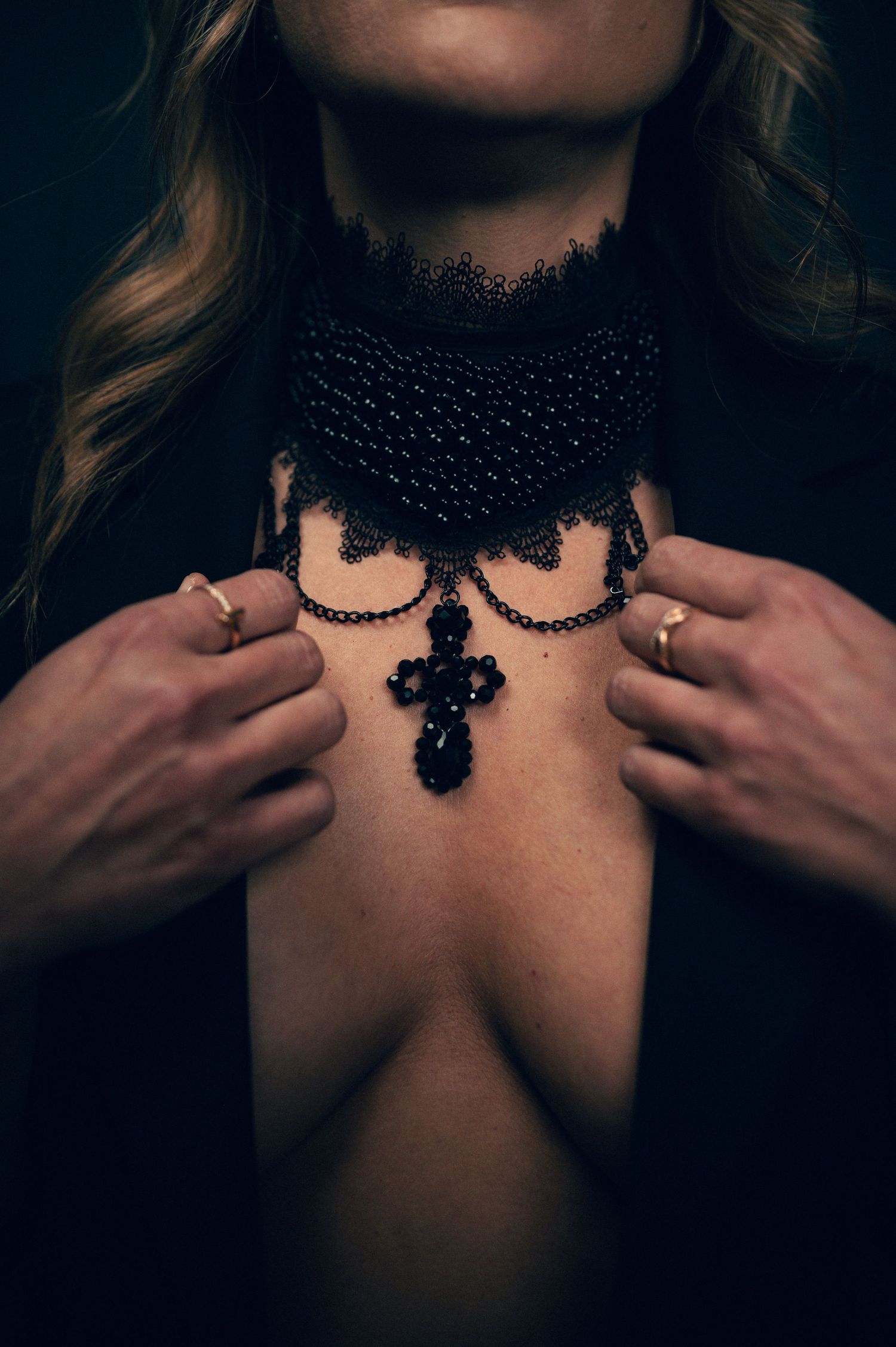 Woman wearing black lace choker necklace with ornate cross pendant, hands adjusting jewelry, dark moody background.