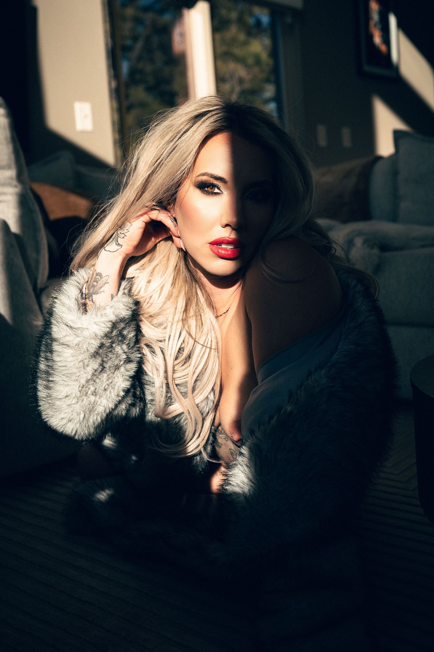 Blonde woman in fur coat with red lips and tattoo poses dramatically in moody indoor lighting.