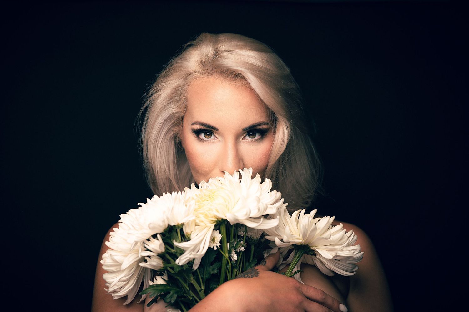 Blonde woman holding white chrysanthemum flowers close to her face against a dark background.