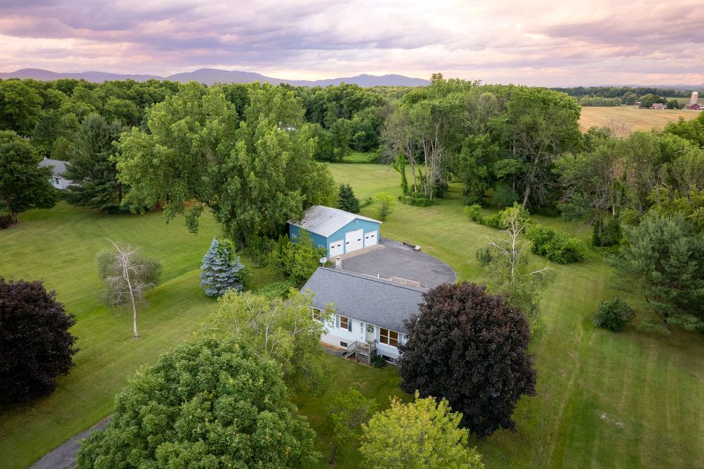 Aerial view of a charming rural home in Washington County surrounded by lush green trees with mountain views and open fields at sunset.