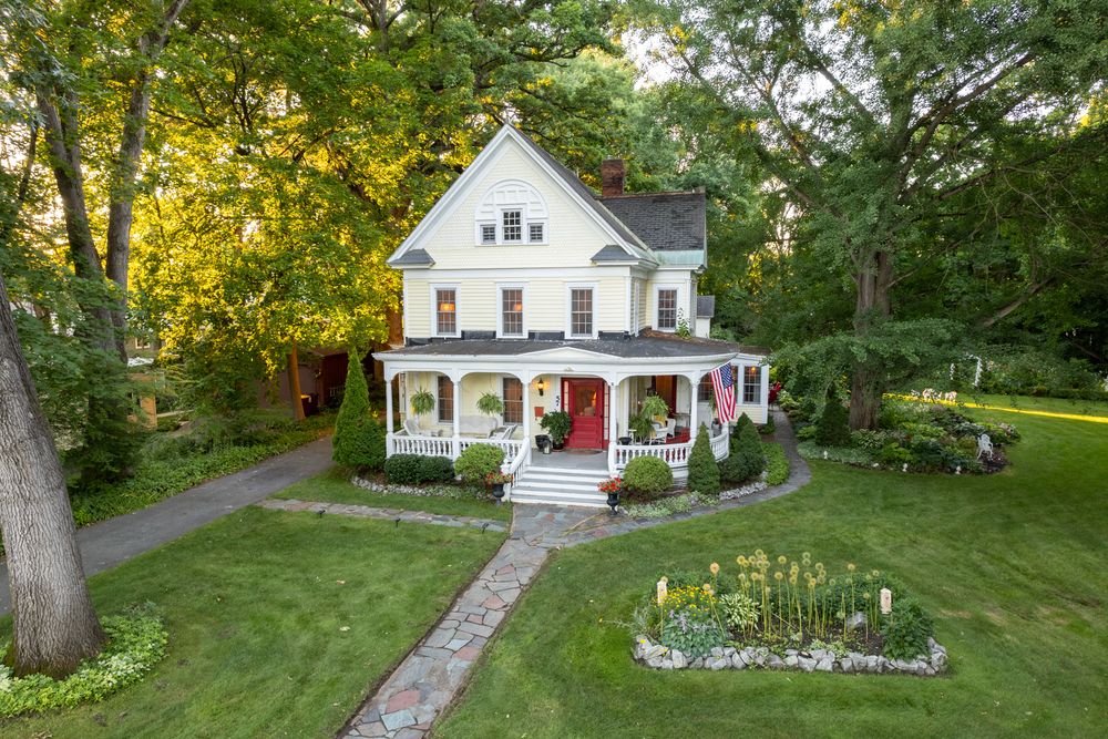 Drone photo of gorgeous Victorian home with wrap-around porch, red door, circular driveway, and lush green lawn surrounded by mature trees.