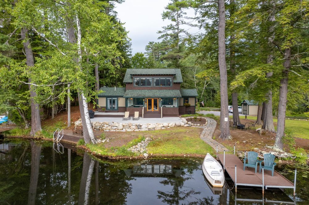 Aerial view over water of a charming lakeside home in Lake Luzerne surrounded by tall pine trees with a private dock and boat.