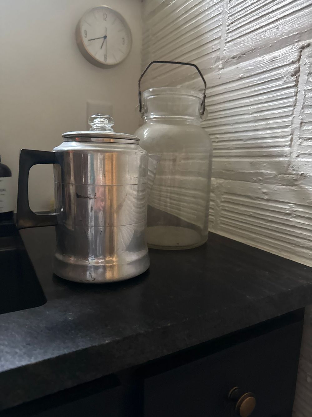Vintage aluminum percolator and glass mason jar on dark granite countertop with textured backsplash and wall clock.