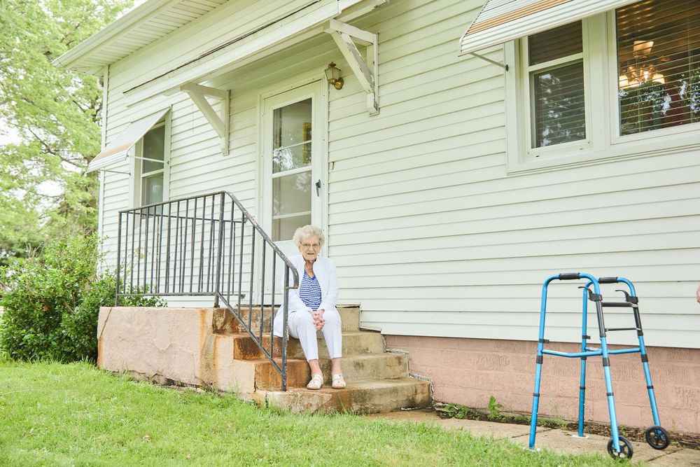 Elderly woman carefully descending front porch steps beside a walker mobility aid outside a light blue house.