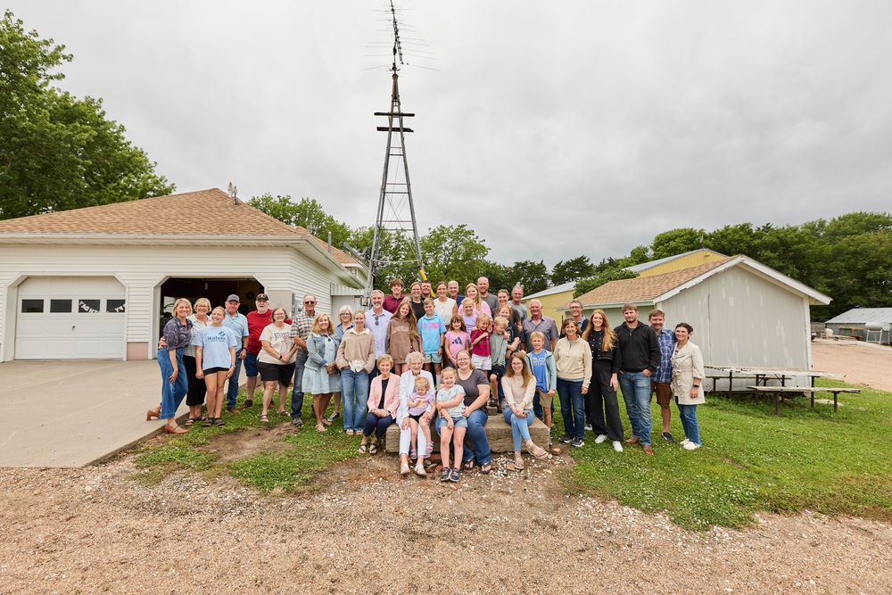 Large family group photo outdoors on a farm with a windmill and white buildings in the background.