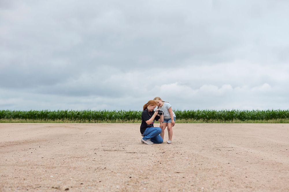 Mother crouching down to embrace young child in open dirt field with green cornfield and cloudy sky.