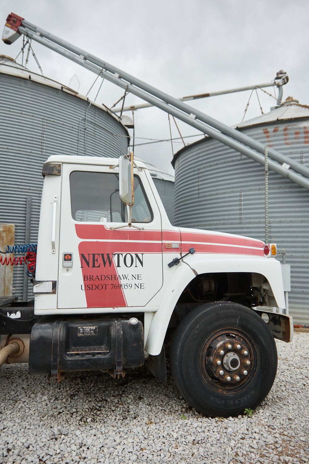 White Newton grain truck from Bradshaw, NE parked beside large metal grain storage silos on a gravel lot.