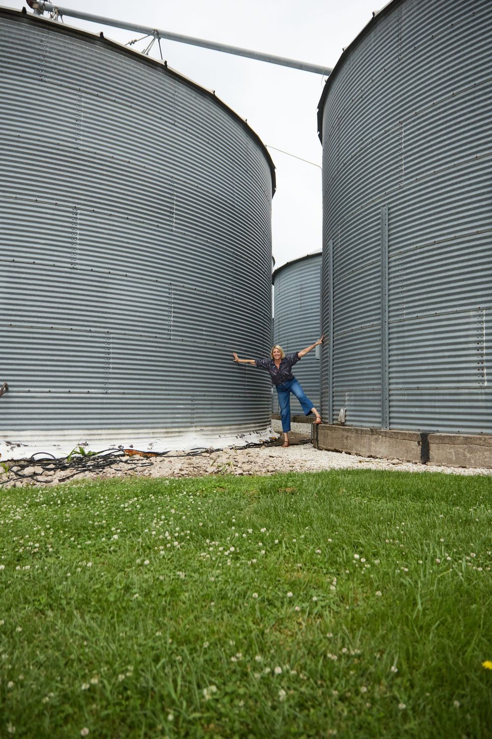Person posing with arms outstretched between large metal grain silos on a farm with green grass.