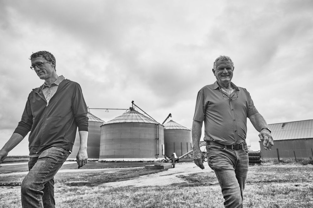 Two farmers walk confidently past grain silos and farm buildings on an overcast day in this black and white rural photo.