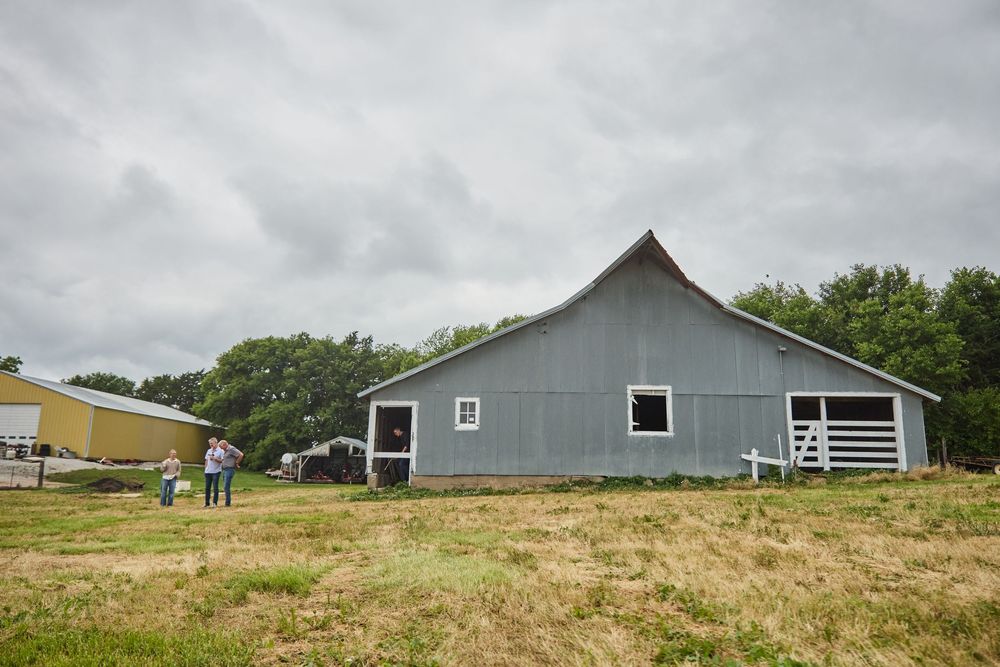 A gray barn stands in an open field under overcast skies on a rural farm property.