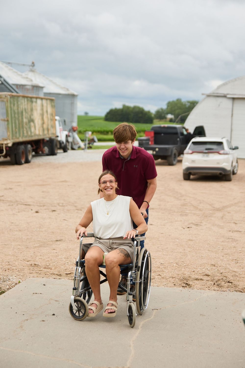 Young man pushing smiling woman in wheelchair on gravel farm path with grain silos and vehicles in background.