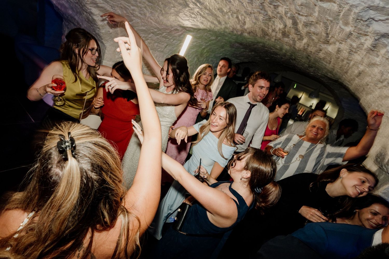 Guests dancing and celebrating at a lively party in a stone vaulted venue, holding drinks and smiling.
