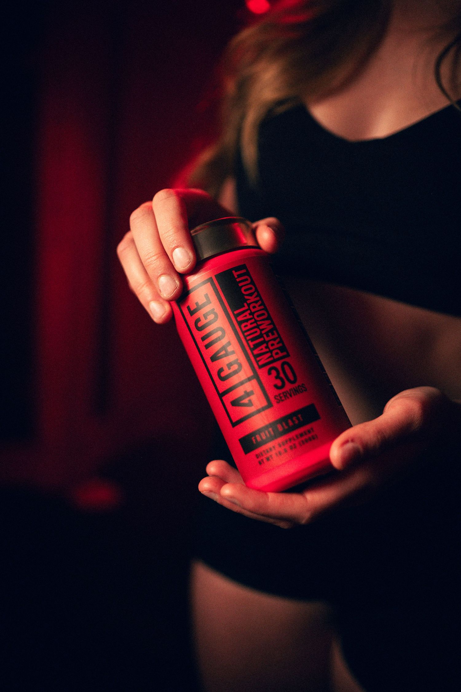 Woman holding a red A1 Gauge pre-workout supplement container in a dark gym setting.
