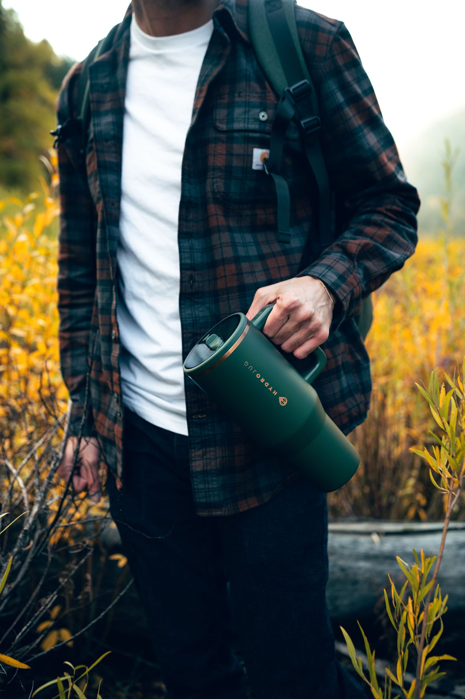 Man in plaid flannel shirt holding a green insulated tumbler outdoors among autumn foliage.