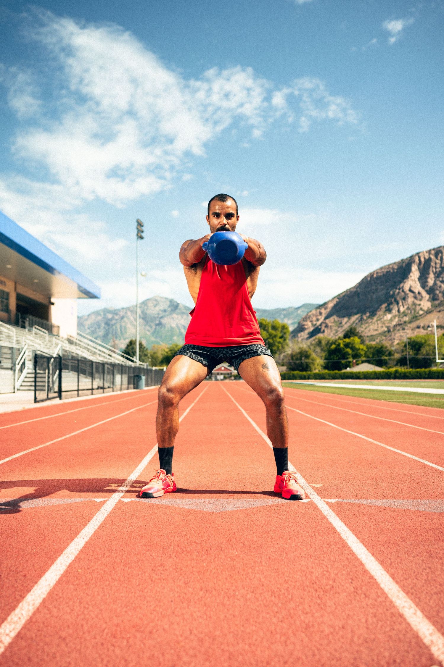 Athletic man in red tank top performs kettlebell squat on outdoor running track with mountains in background.