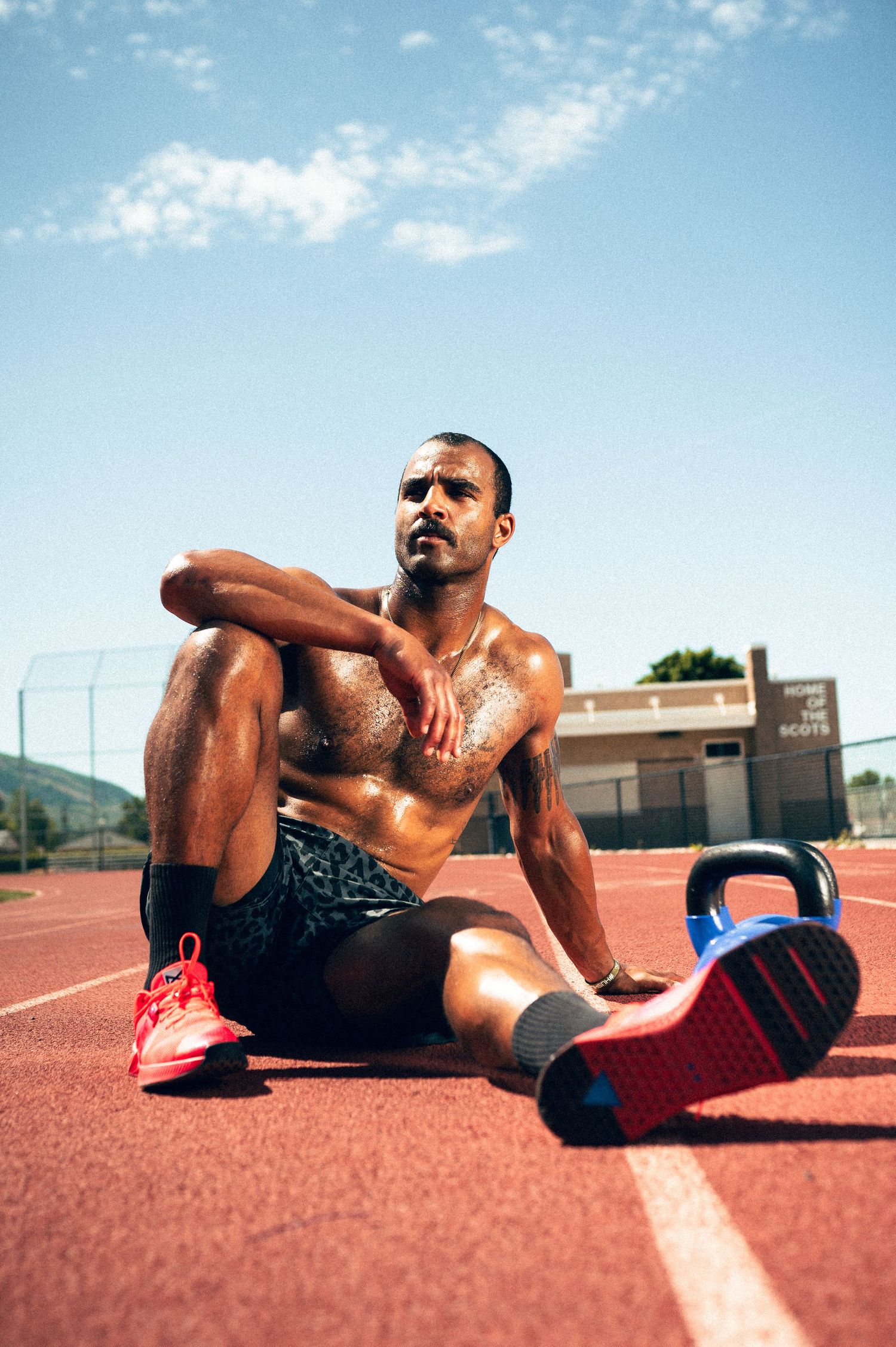 Muscular athlete resting on red track after intense outdoor workout with kettlebell nearby.
