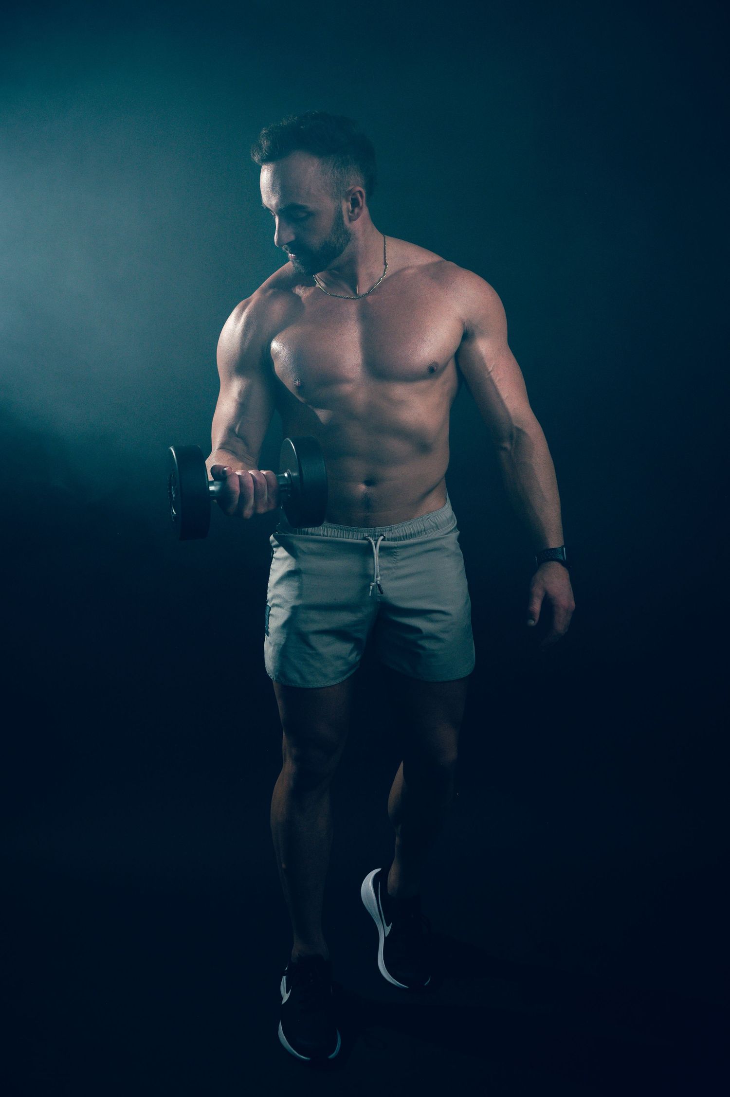 Muscular shirtless man curling a dumbbell in a dark gym, showcasing defined abs and biceps in gray shorts.