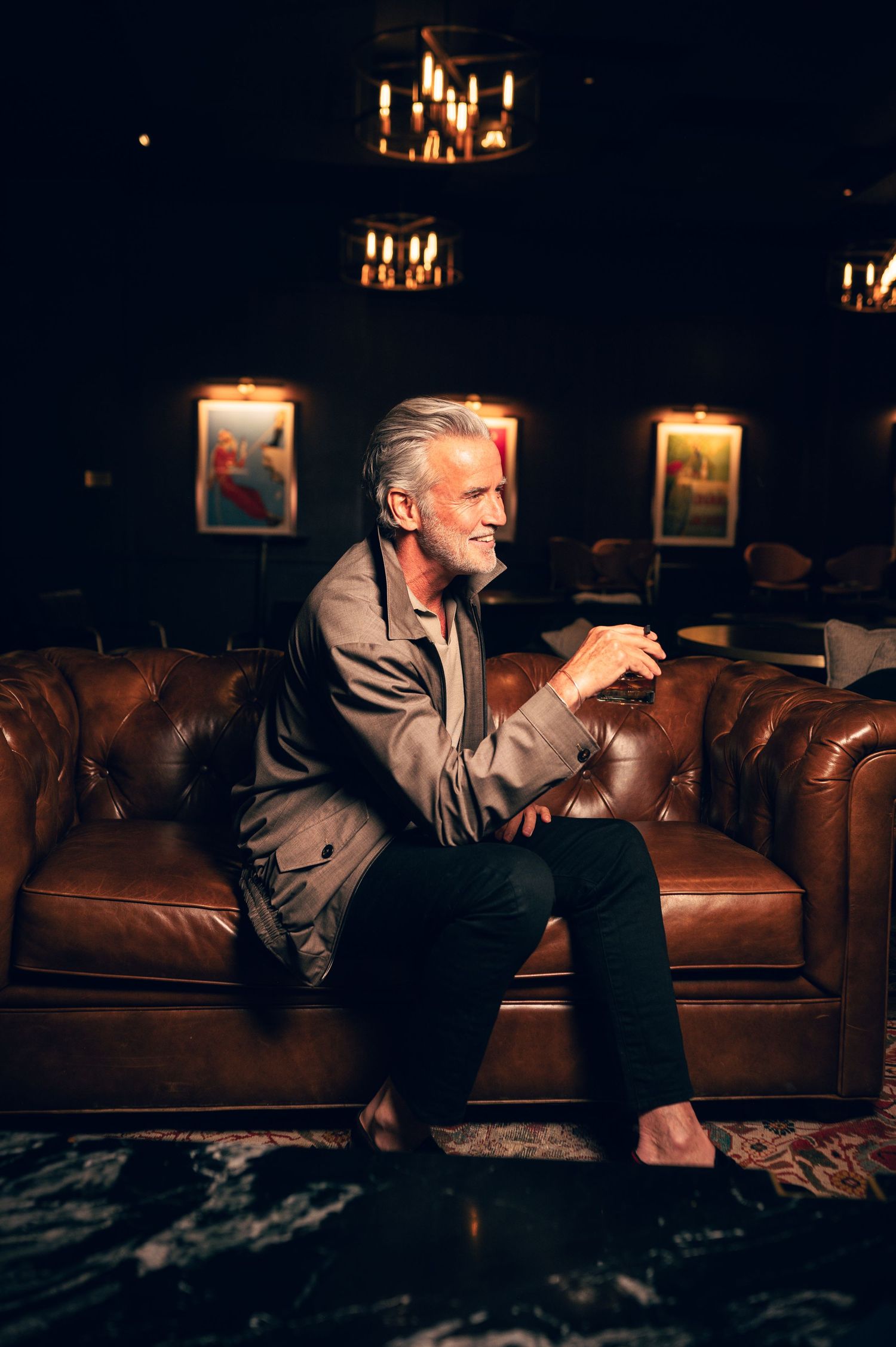 Distinguished silver-haired man sitting on a leather Chesterfield sofa in a dimly lit upscale lounge.