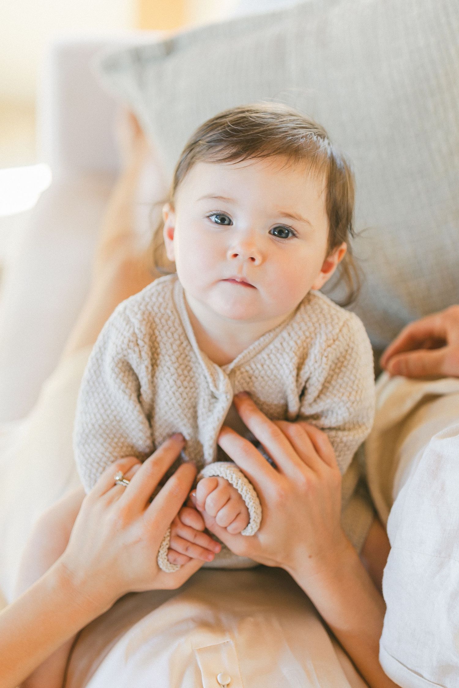 Adorable baby in beige knit sweater sitting on couch, held gently by mother's hands, looking up with blue eyes.
