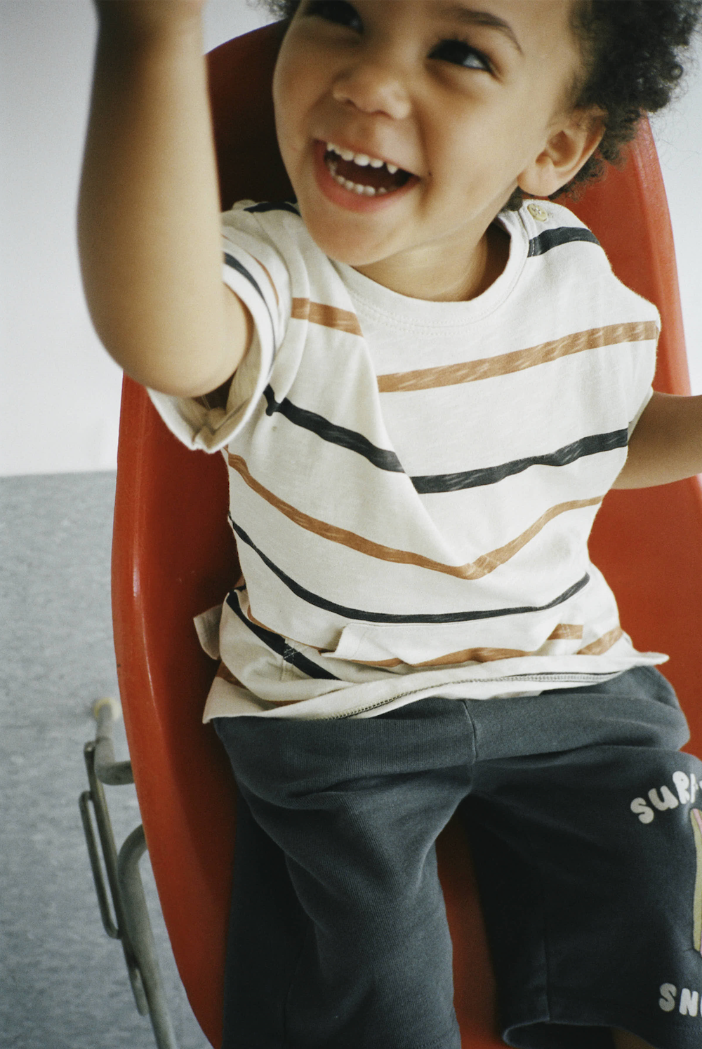 Happy toddler in striped white tee and dark joggers laughs while sitting in red chair.