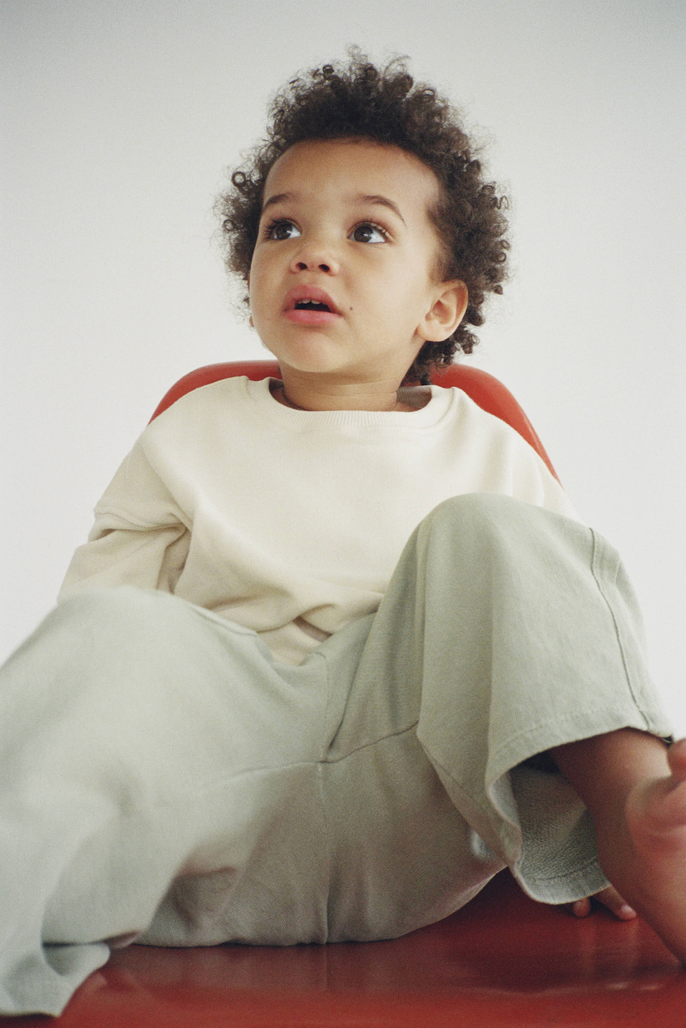 Curious toddler in cream sweatshirt and wide-leg pants sits on red chair looking upward.
