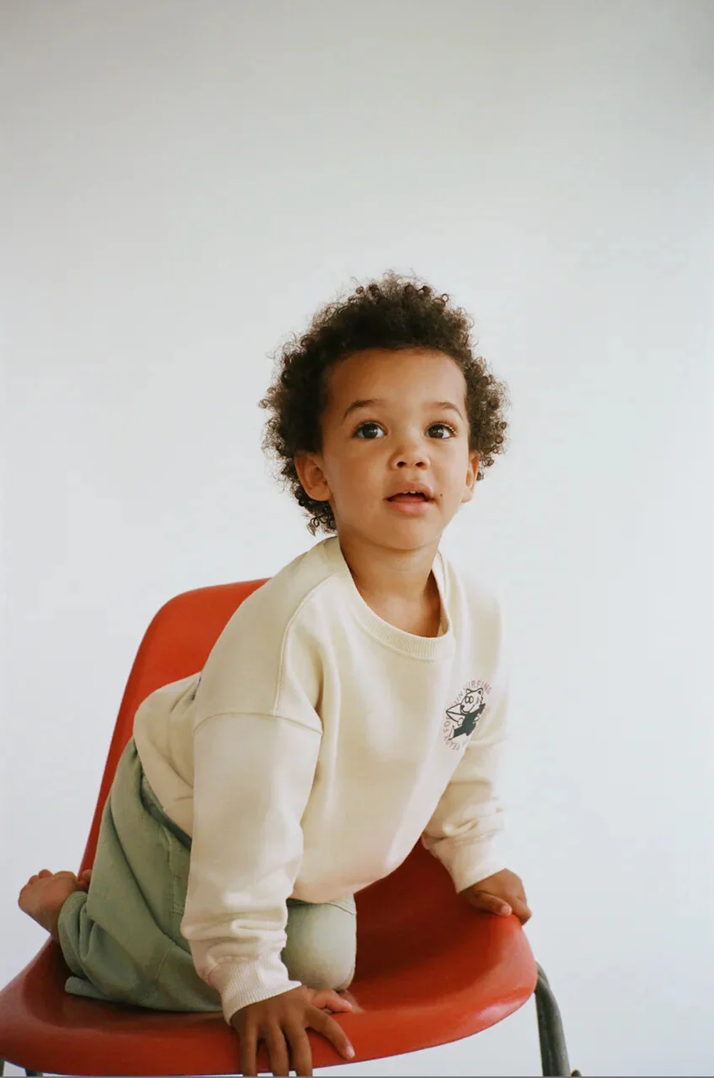 Young child in cream sweatshirt and green pants sits on red chair with thoughtful expression.