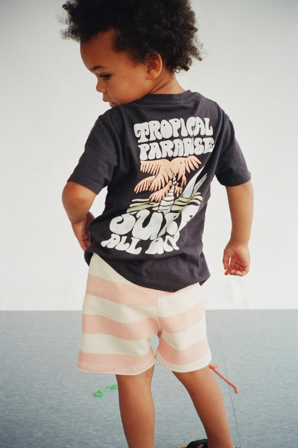 Toddler in dark graphic tee and pink striped shorts walks away showing Tropical Paradise print.