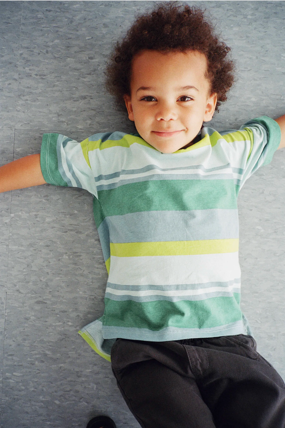 Smiling boy in green striped t-shirt and black pants lies on gray floor looking up.
