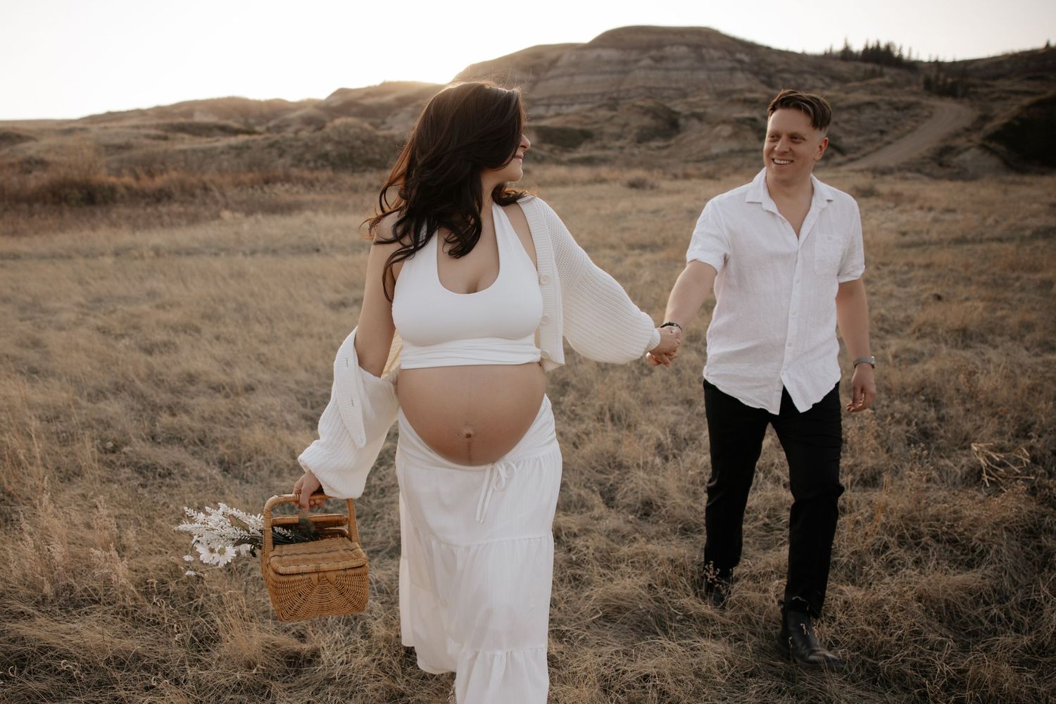 Pregnant couple holding hands walking through lush green hillside meadow at golden hour in flowing white maternity dress.