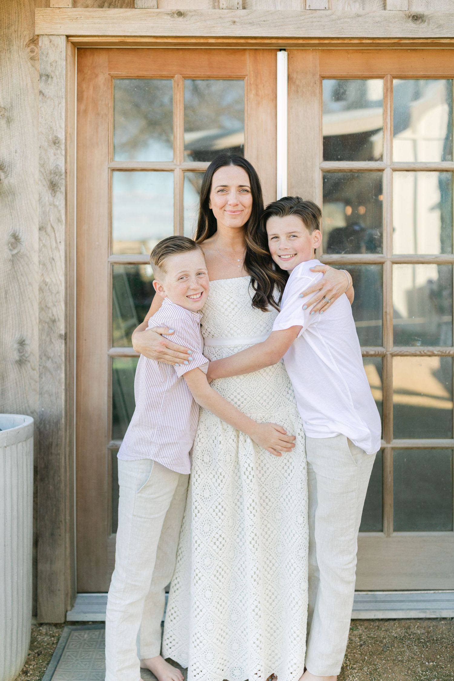 Couple embracing in front of rustic wooden barn doors, woman in white lace dress, man in light shirt and gray pants.