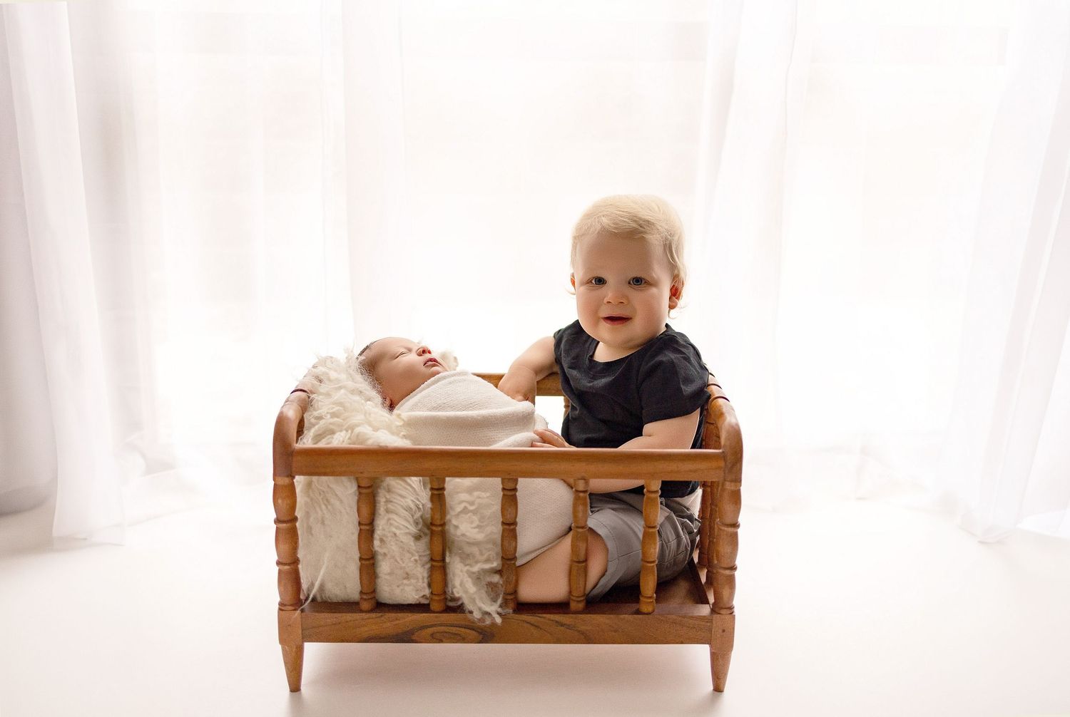 Toddler boy stands beside a wooden doll bed holding a swaddled newborn baby in a bright white studio.