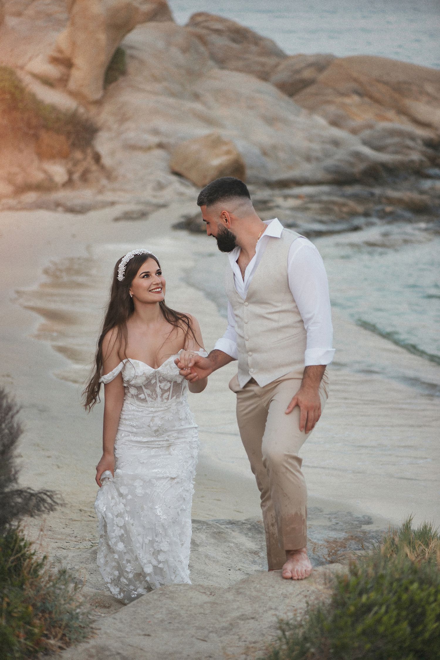 Candid wedding photography of a bride and groom walking hand-in-hand on a rocky beach in Greece.