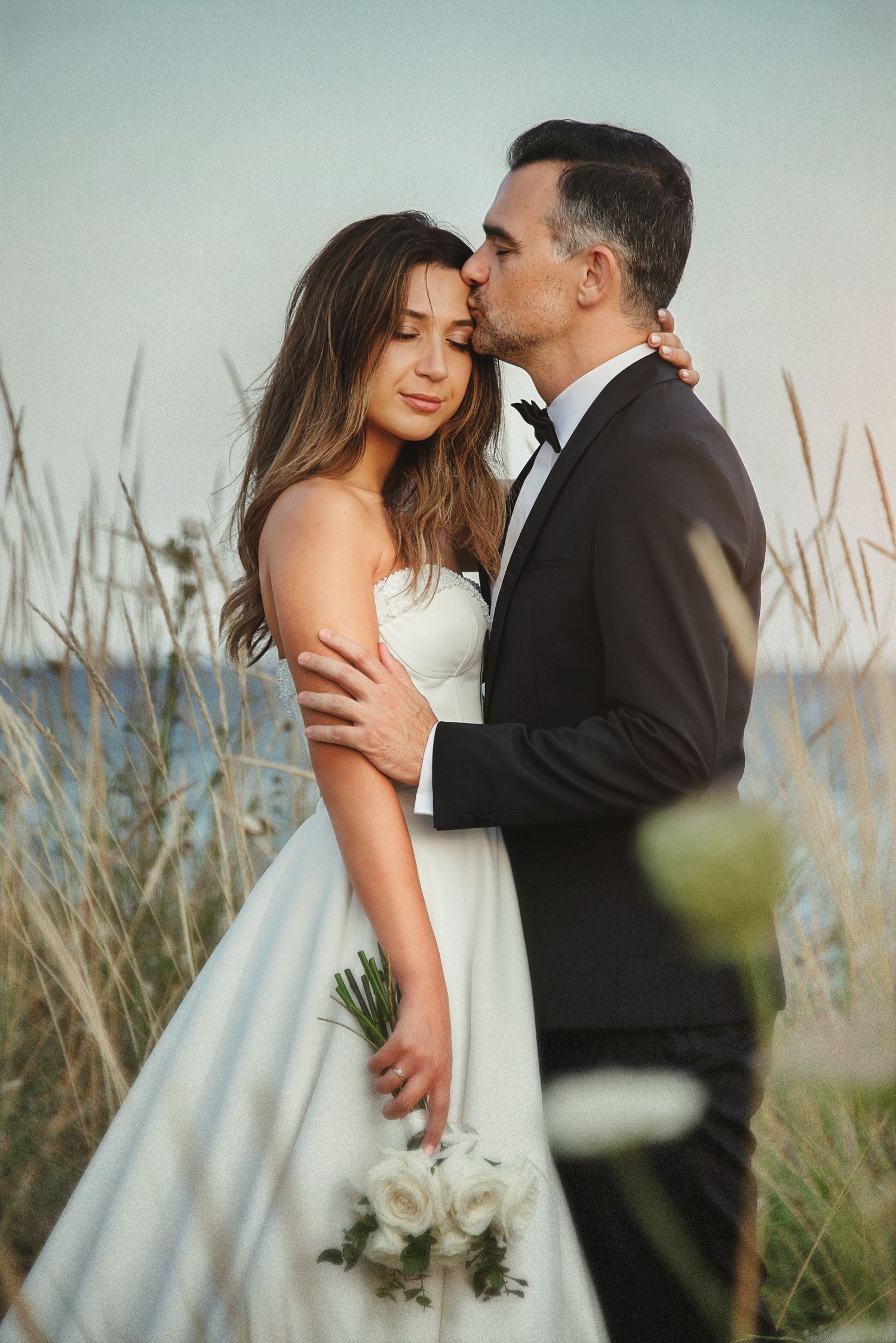 Candid coastal wedding photography featuring a groom kissing the bride's forehead in a natural tall grass setting.