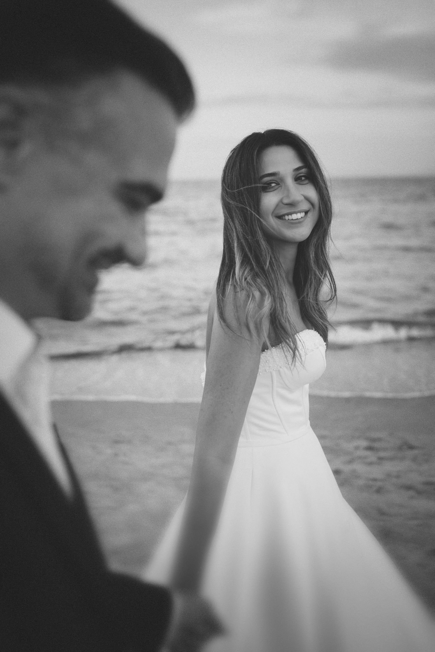 Candid black and white wedding photography featuring a smiling bride walking hand-in-hand with the groom on a beach in Greece.