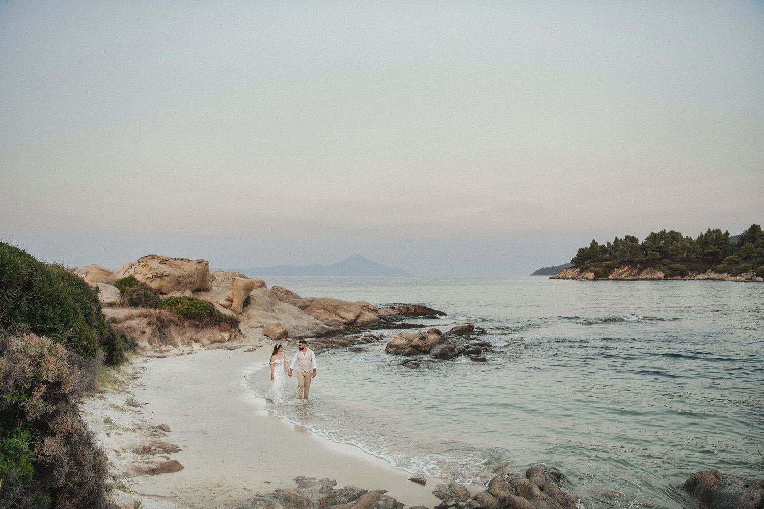 Cinematic destination wedding photography, bride and groom walking on a rocky beach in Greece during sunset.