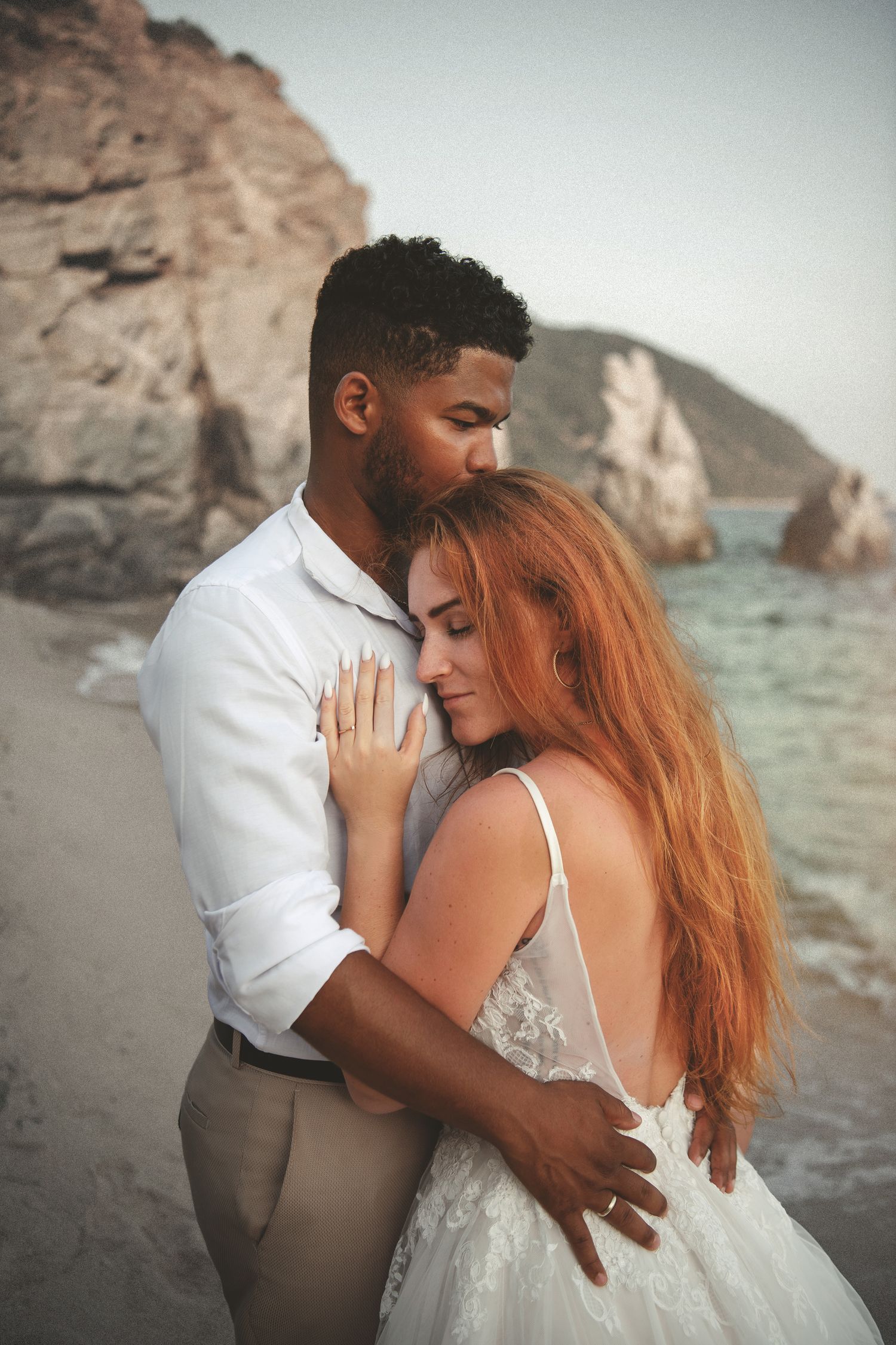 Candid destination beach wedding photography featuring an intimate bride and groom embrace by coastal cliffs.