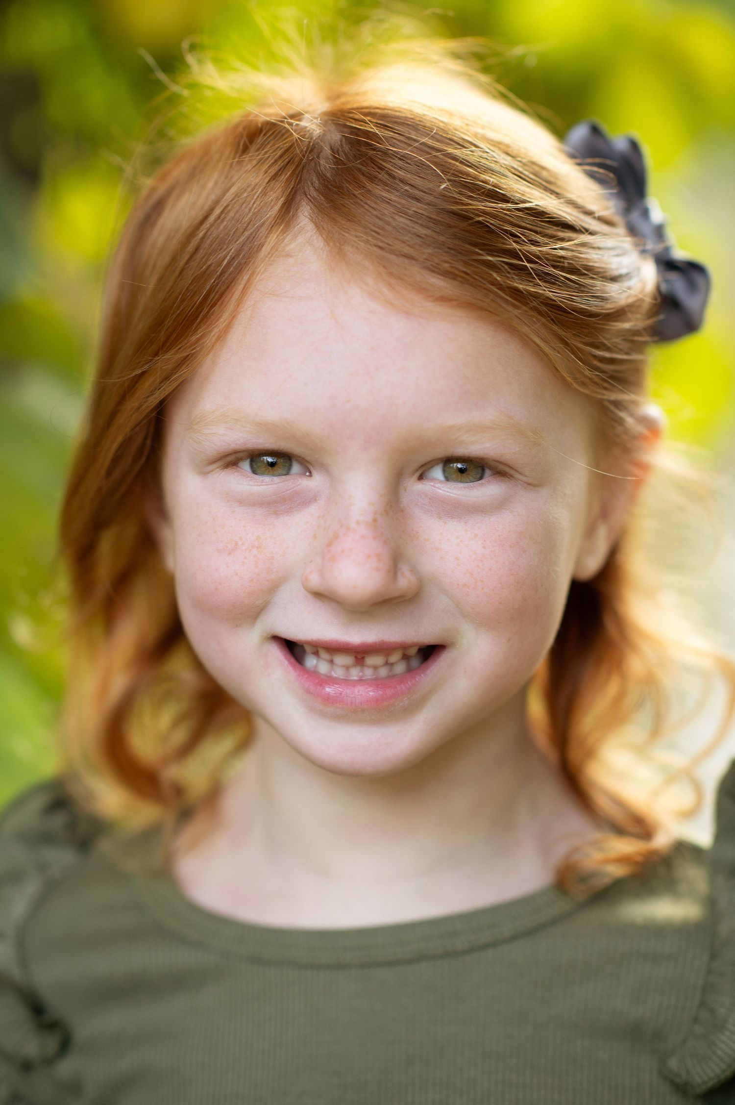 Smiling redhead girl with freckles wearing green top and black bow outdoors.