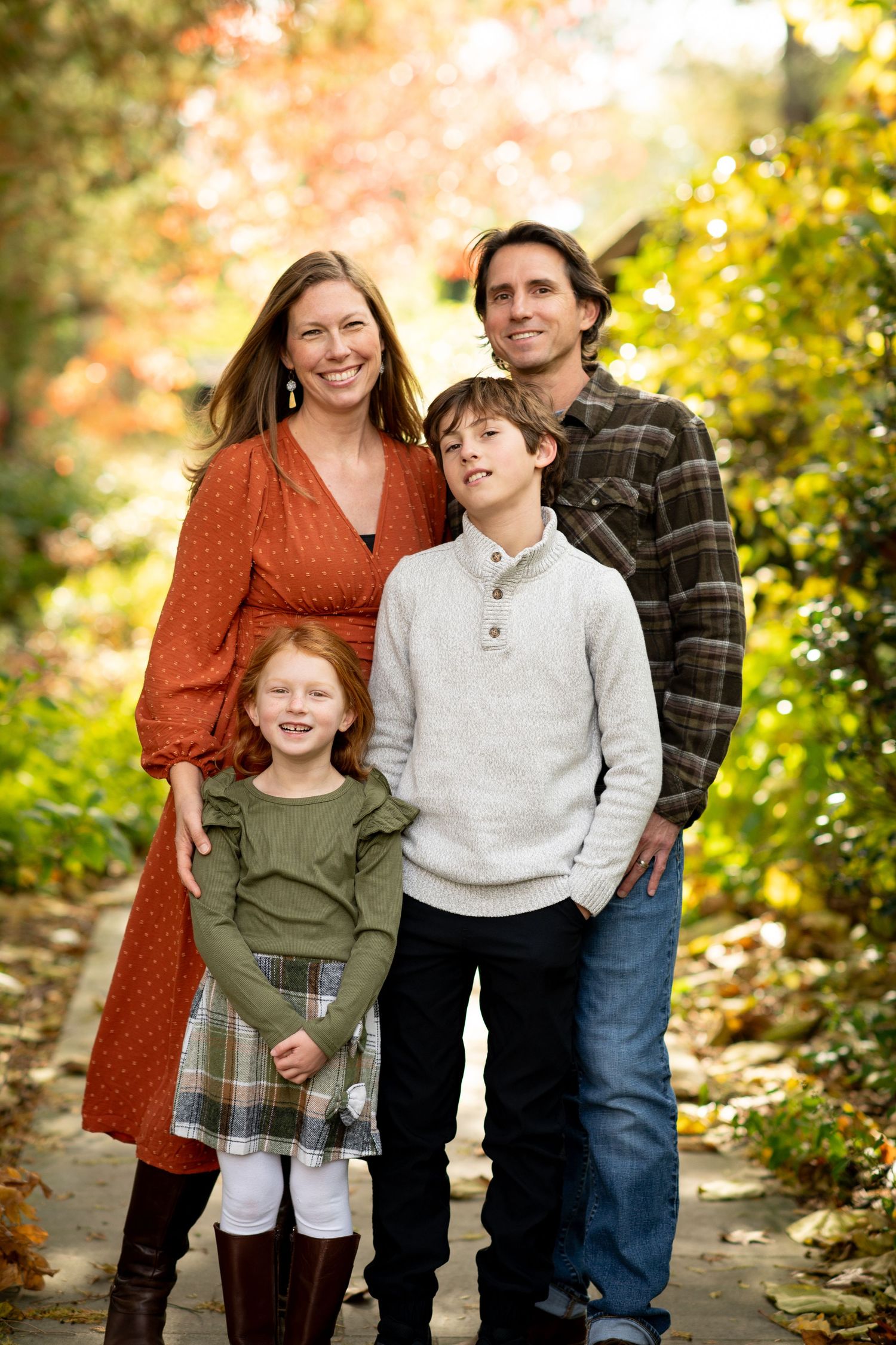 Family of four posing outdoors on a path surrounded by colorful autumn foliage, smiling together.