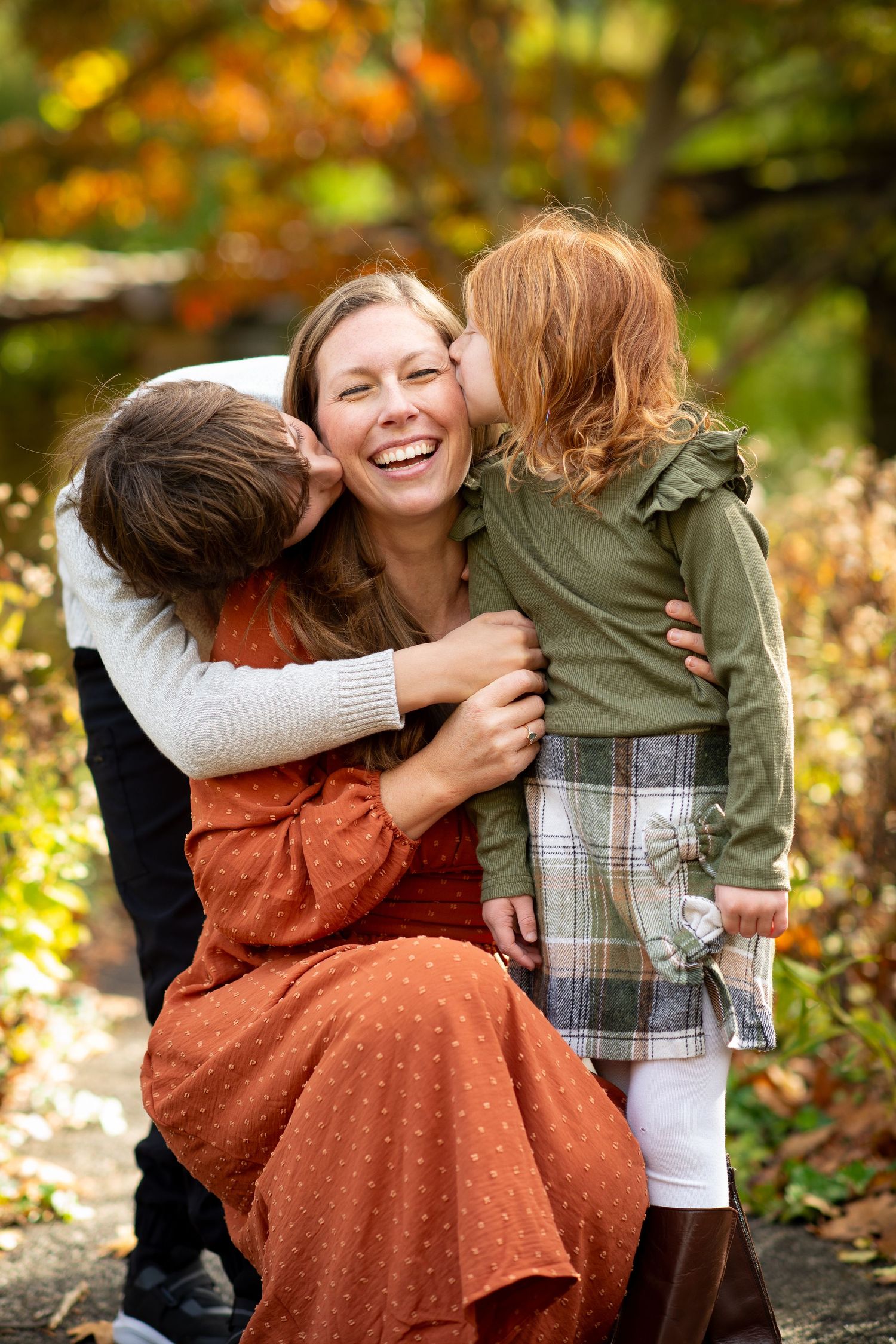 Mother laughing as two children kiss her cheeks in autumn park setting.