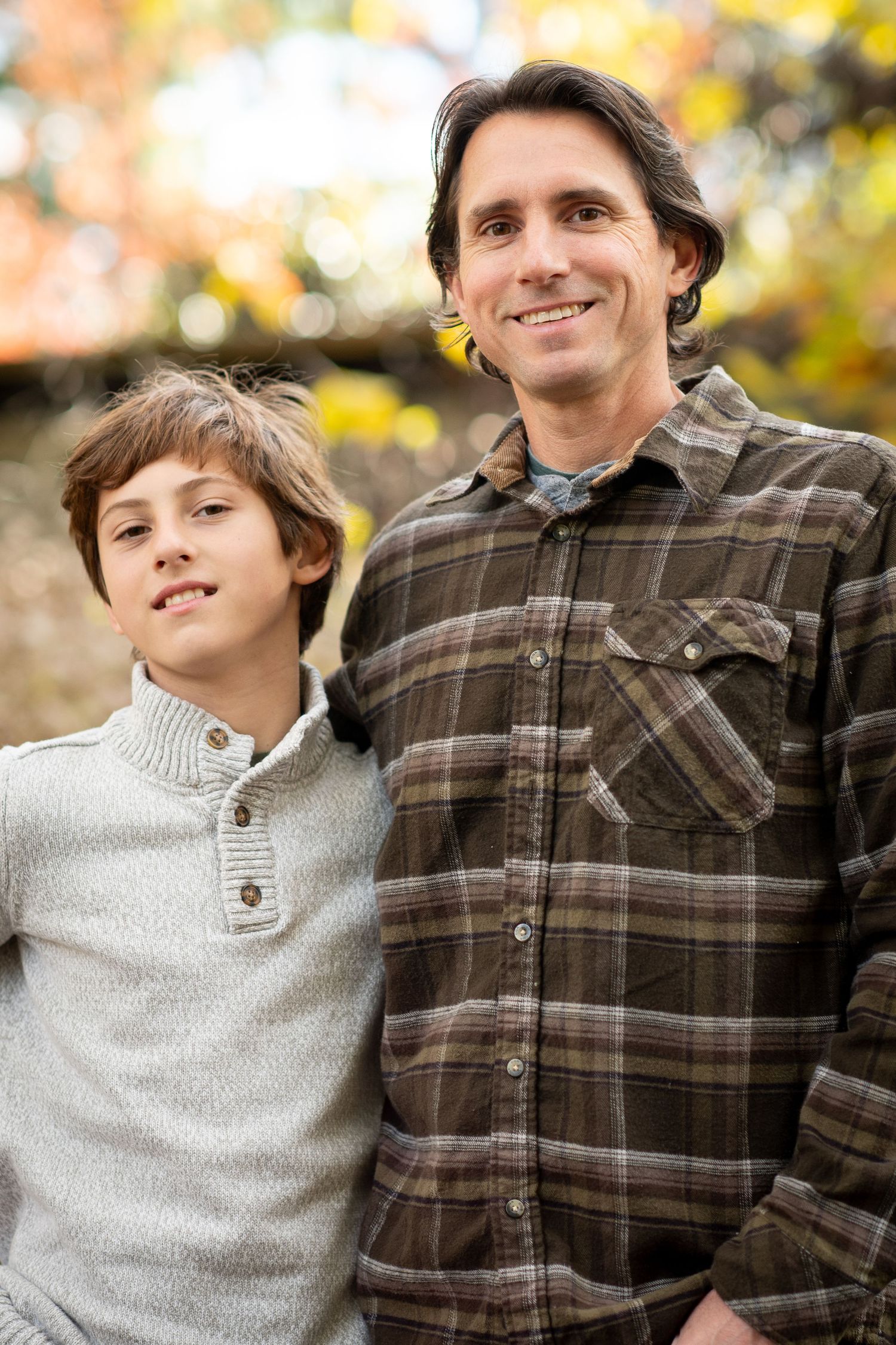 A smiling man and young boy pose together outdoors in autumn, wearing casual sweaters with colorful fall foliage behind them.