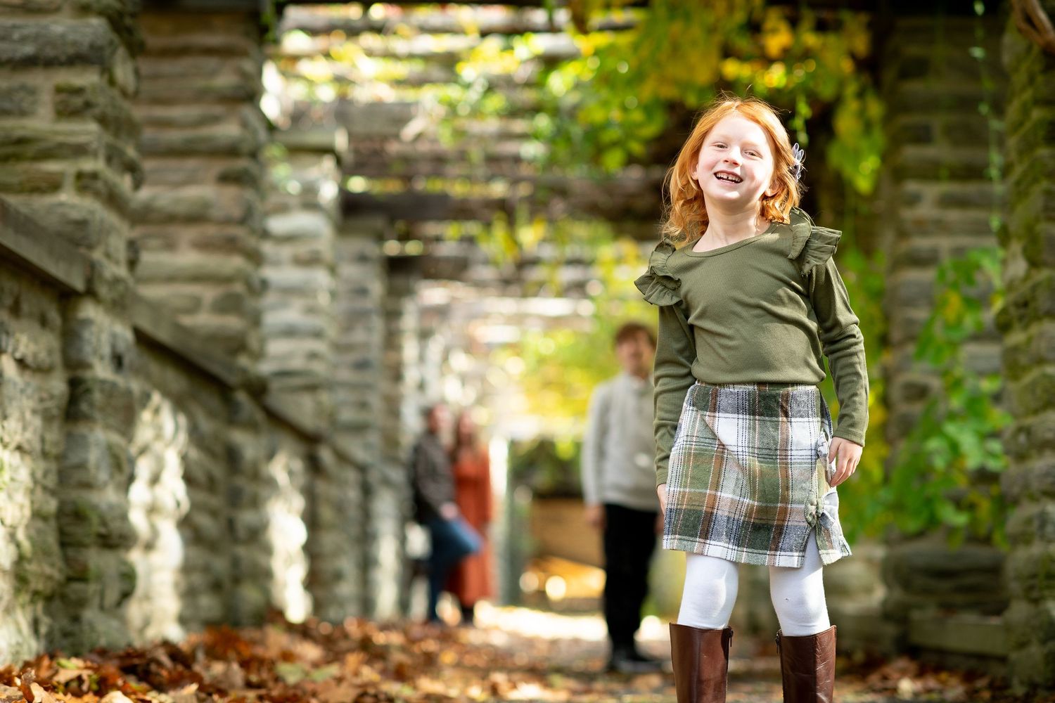 A joyful redhead girl in a plaid skirt and green top laughs outdoors among stone columns with autumn foliage.