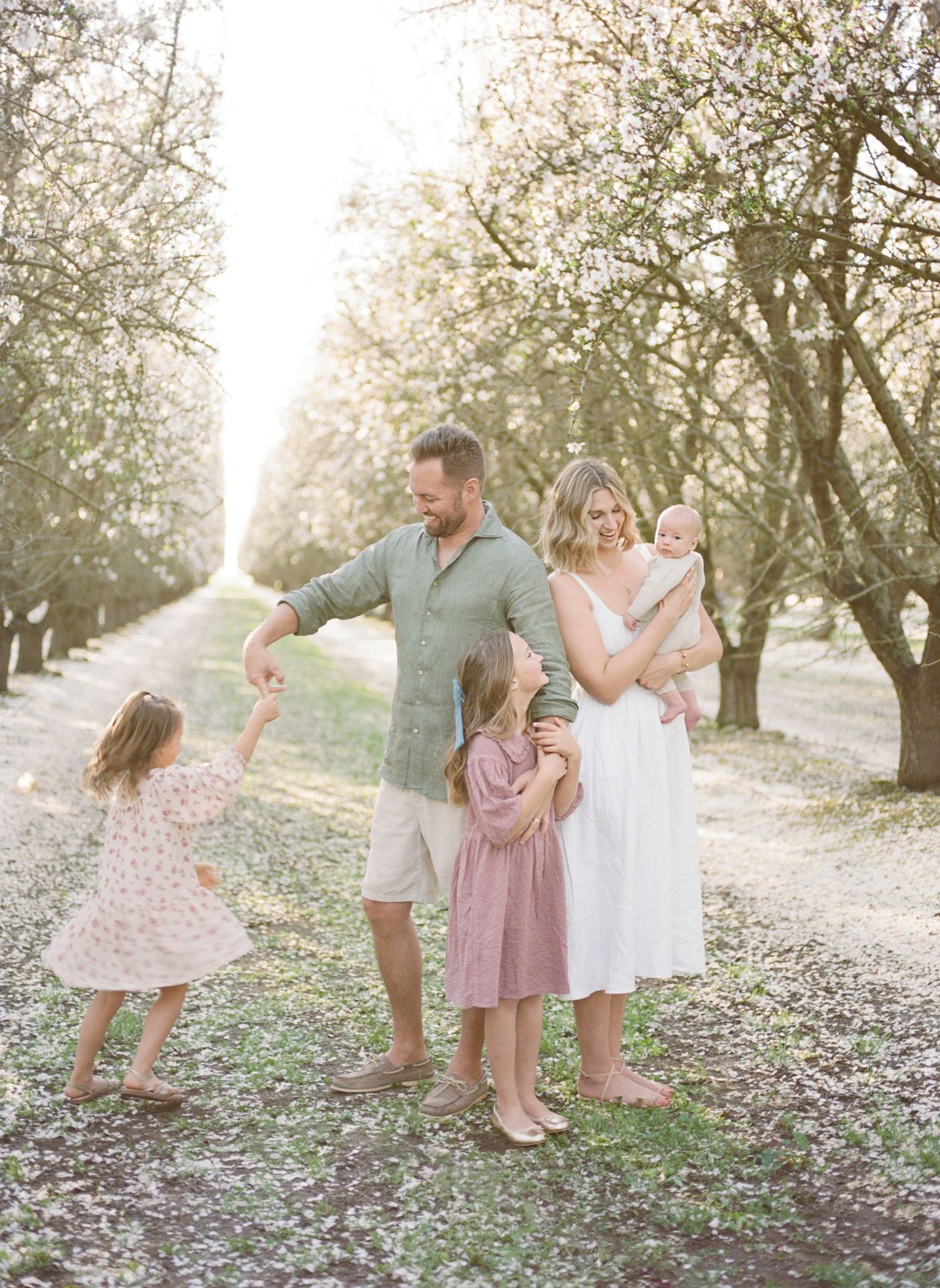 Family of five enjoying a sunny spring day among blooming trees, with children in pastel dresses and parents smiling warmly.