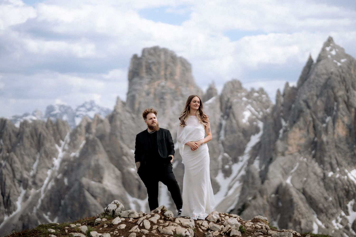 Couple in wedding attire posing on rocky mountain peak with dramatic Dolomites peaks and clouds in background.