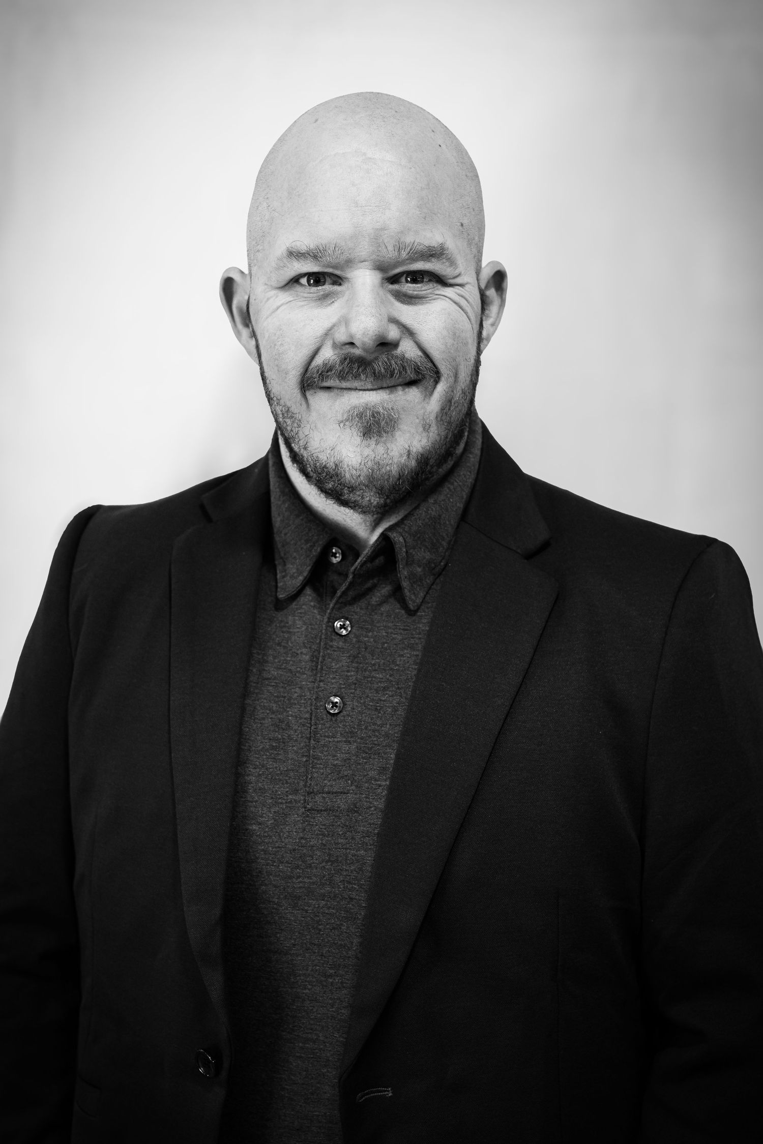 Bald man with beard and mustache wearing black blazer over gray shirt, smiling in professional black and white portrait.