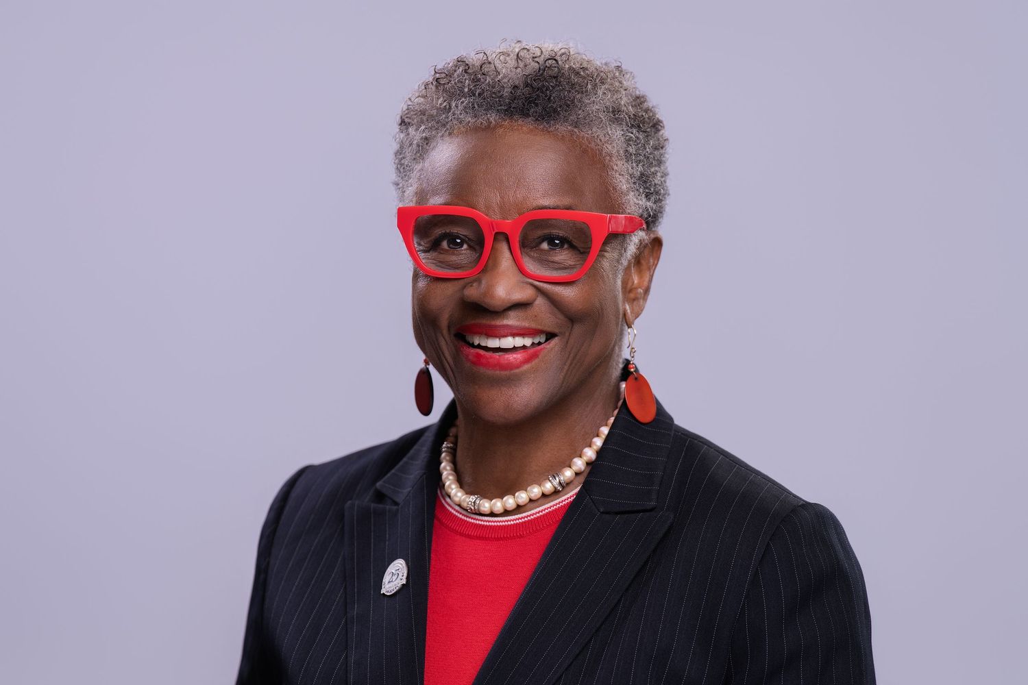 Professional headshot of a smiling woman wearing red glasses, red top, pearl necklace, and black blazer against gray background.