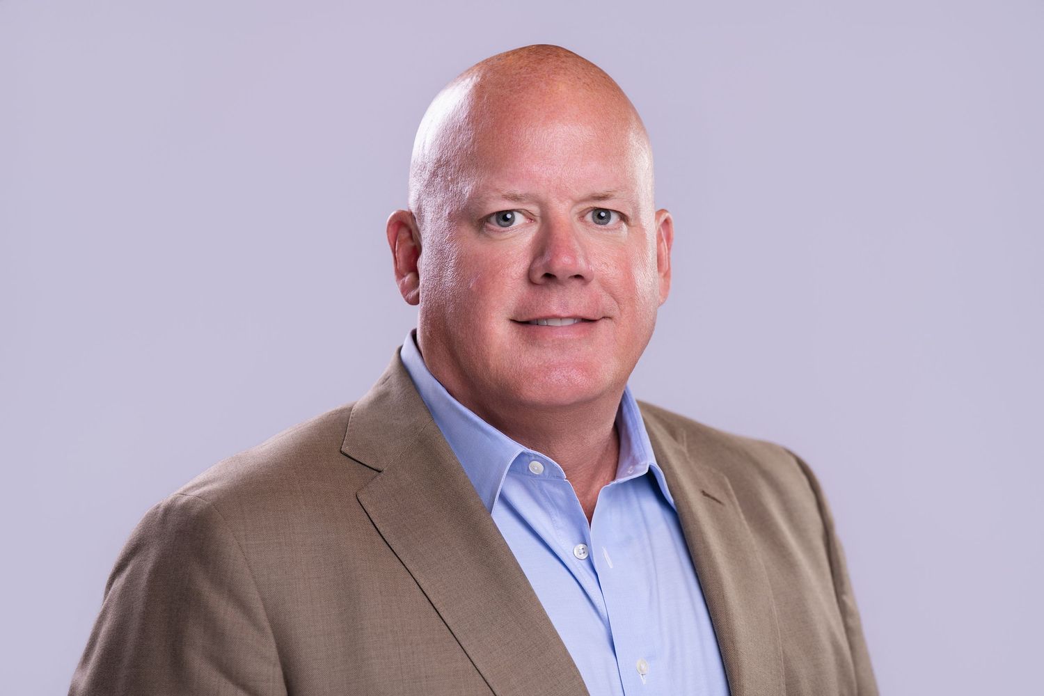 Professional headshot of a smiling businessman in a tan blazer and blue shirt against a gray background.