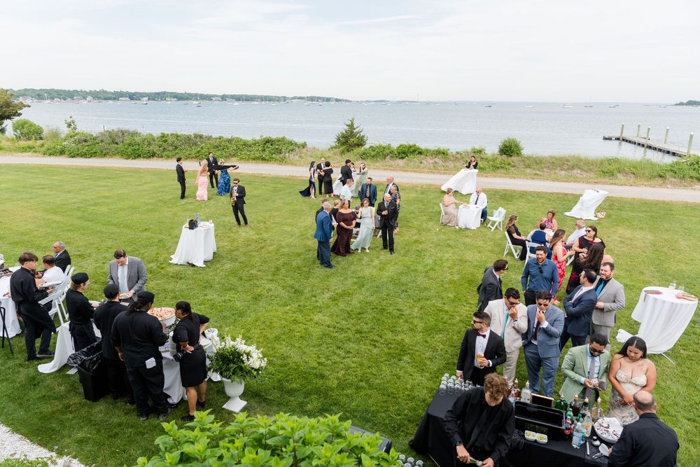 Aerial view of an elegant outdoor wedding reception on a green lawn overlooking a serene waterfront.